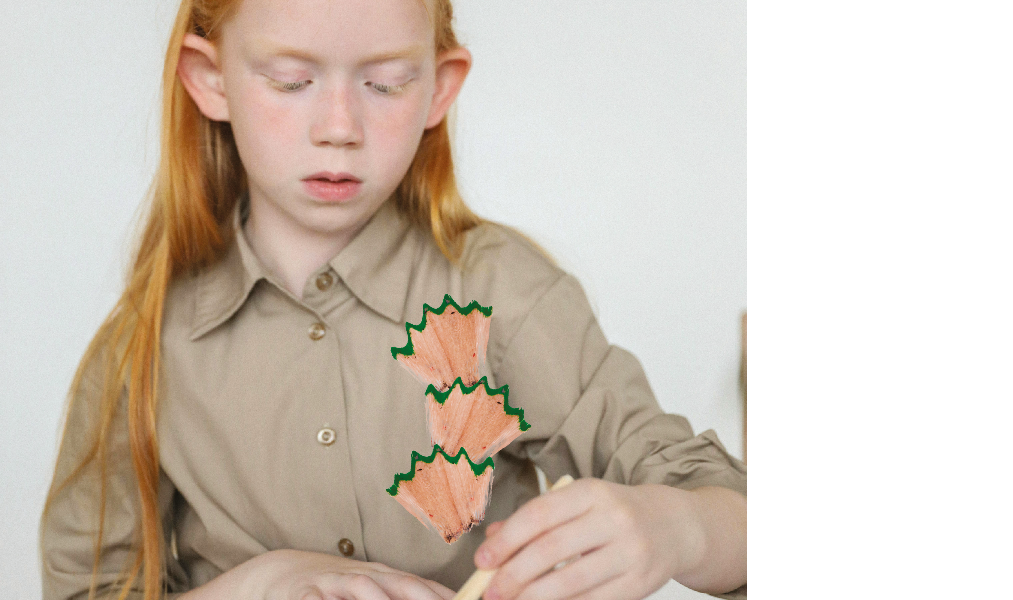 Ein Kind mit langen roten Haaren arbeitet konzentriert mit den Händen an einem Objekt, dekoriert mit Bleistiftspänen.